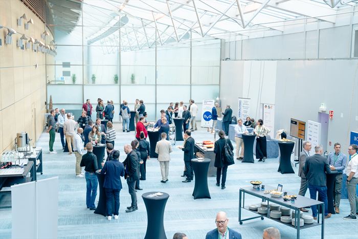 Espace de networking sous la verrière : participants autour des mange-debout, stands des partenaires en fond